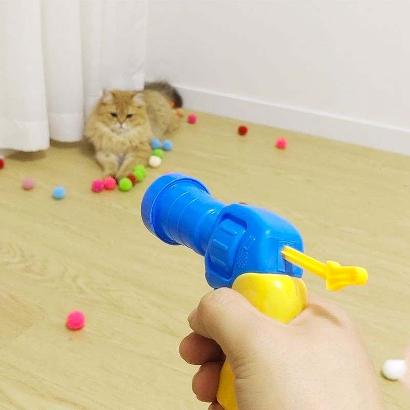 Automatic Ball Launcher toy being aimed at a cat with colorful balls scattered on a wooden floor