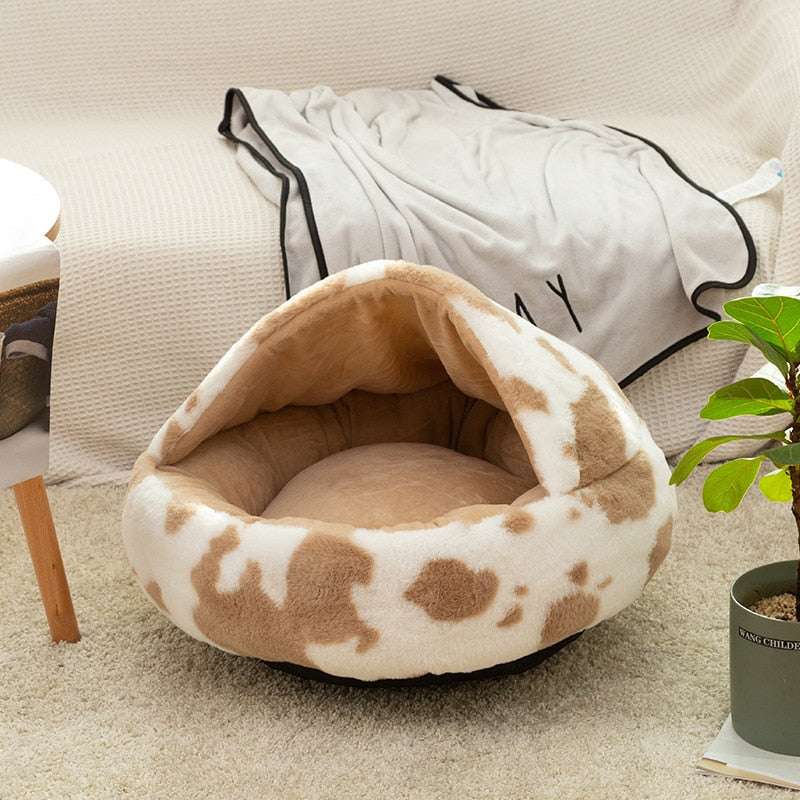 Plush pet cat bed in beige and white cow pattern with soft, cozy fabric and cave-like design on carpeted floor