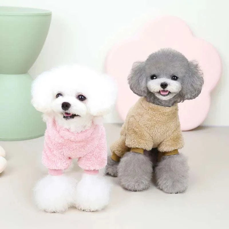 Two small dogs wearing soft fleece elastic pet jumpsuits, one in pink and one in beige, sitting indoors on a light floor with pastel background.